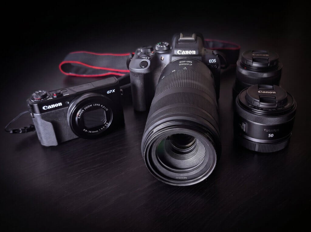 My photography gear journey with a lightweight mirrorless travel camera setup featuring a Canon EOS R10 with a long telephoto zoom lens, a compact Canon PowerShot G7 X pocket camera, a 50mm prime lens, and a 18-45mm wide angle lens on a dark textured surface.