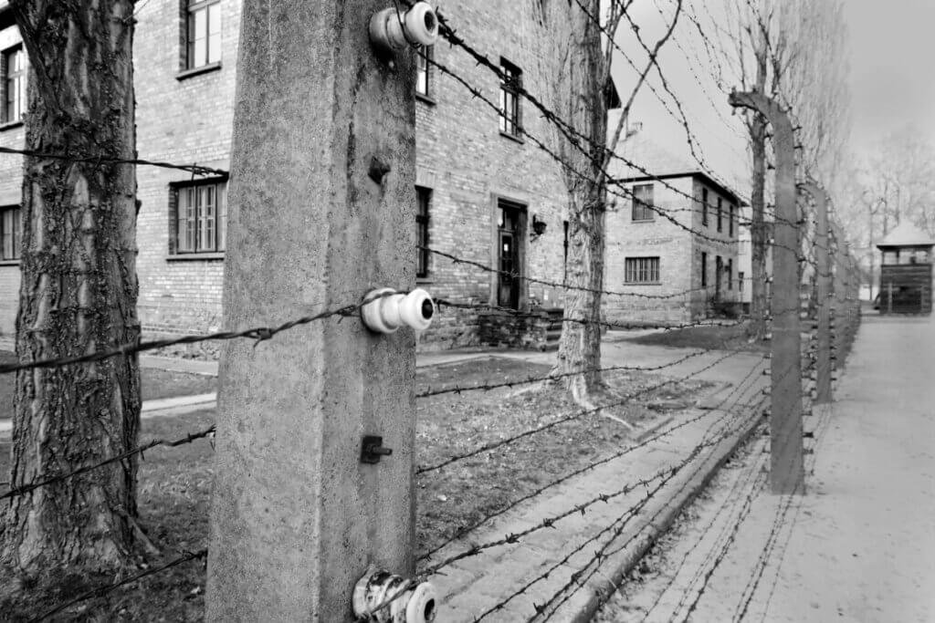 Black and white photograph of double barbed wire fences and brick barracks at Auschwitz I concentration camp.