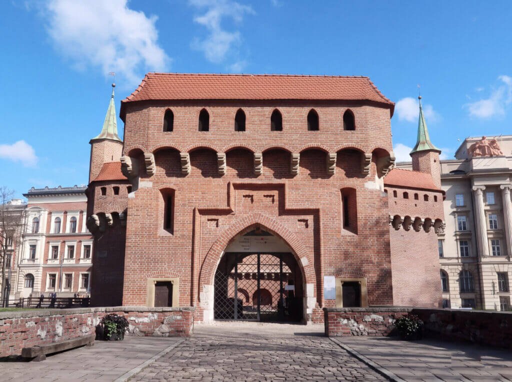 The historic red brick Kraków Barbican fortress featuring a large central pointed arch gateway and small circular watchtowers with green conical spires under a blue sky.