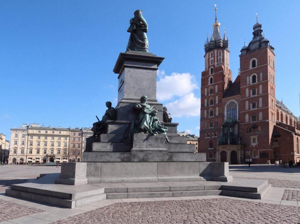 The Adam Mickiewicz Monument standing in the Main Market Square of Kraków, Poland, with the distinct towers of St. Mary's Basilica against a clear blue sky.