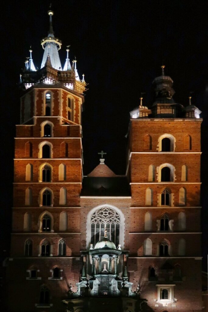 St. Mary's Basilica in Kraków, Poland, beautifully illuminated at night, showing the detailed architecture of its two asymmetrical towers against a dark sky.